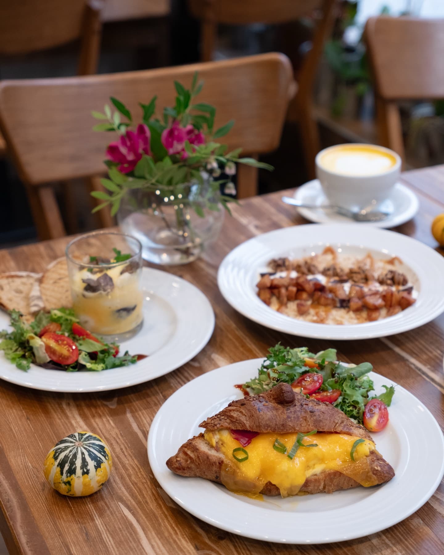 Brunch on a wooden table: croissant with eggs and cheddar, salad, a latte and flowers in a vase